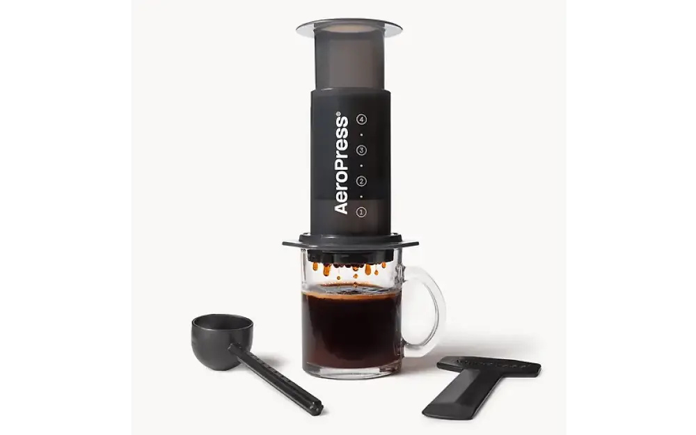 Black aeropress kahvinvalmistuslaite läpinäkyvällä mäntällä ja mittamerkinnöillä varustetulla lasimukilla