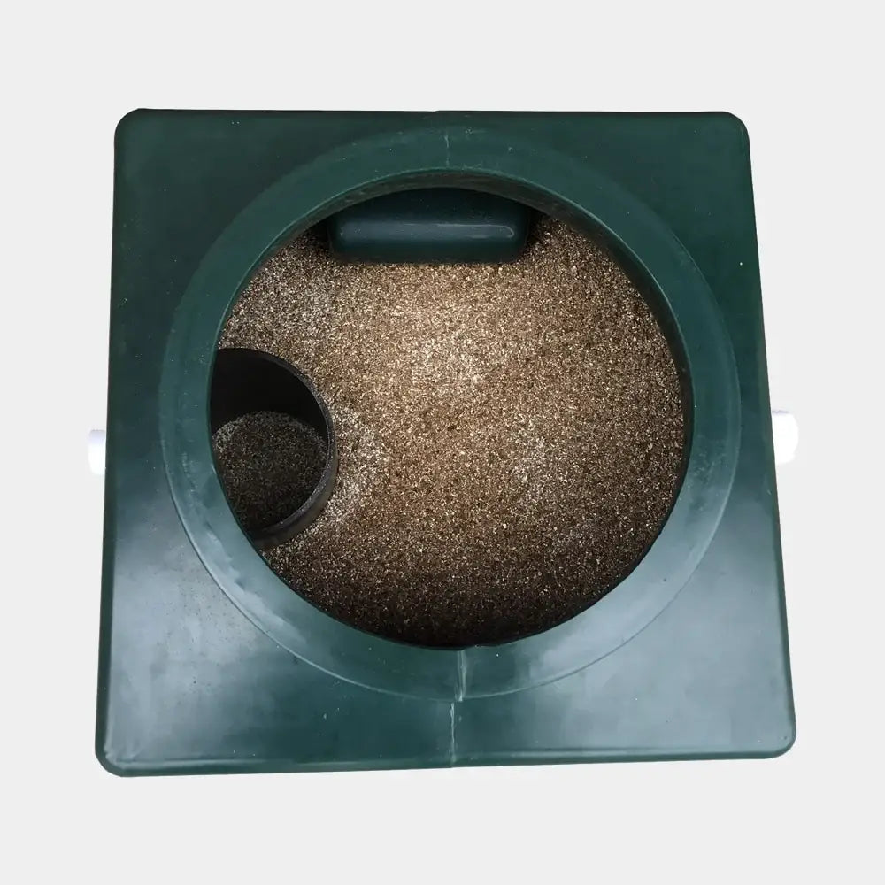 Dark green square plastic geoTrap huussisuodatin container with circular opening filled with brown granular material and small cylindrical component inside