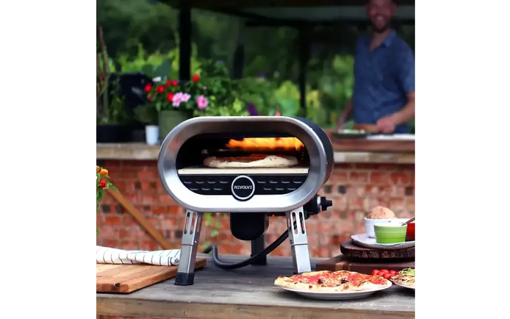 Revolve pizzauuni kaasukäyttöinen hopean ja mustan värinen pyörivä uuni ovaalin muotoisilla tukevilla metallijaloilla ja pizzalla sisällä