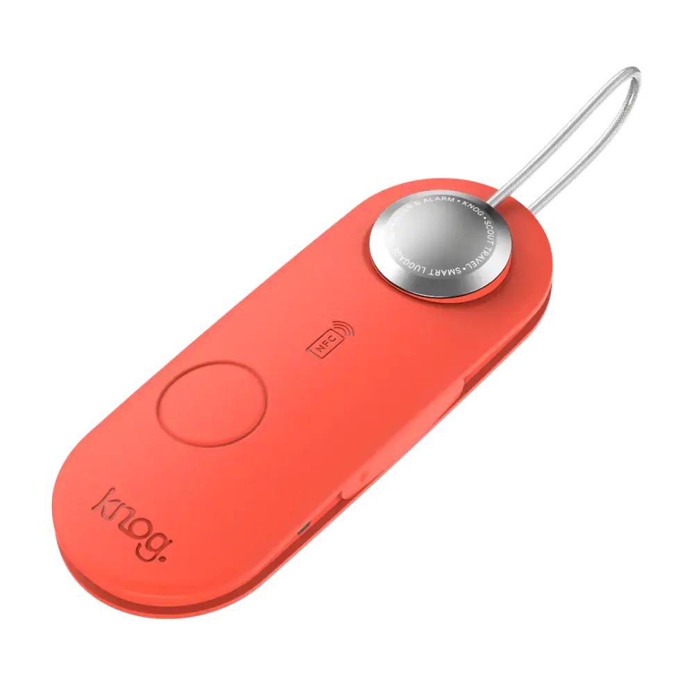 A sleek coral-colored knog smart luggage alarm with a silver button and metallic keychain loop in scout travel red