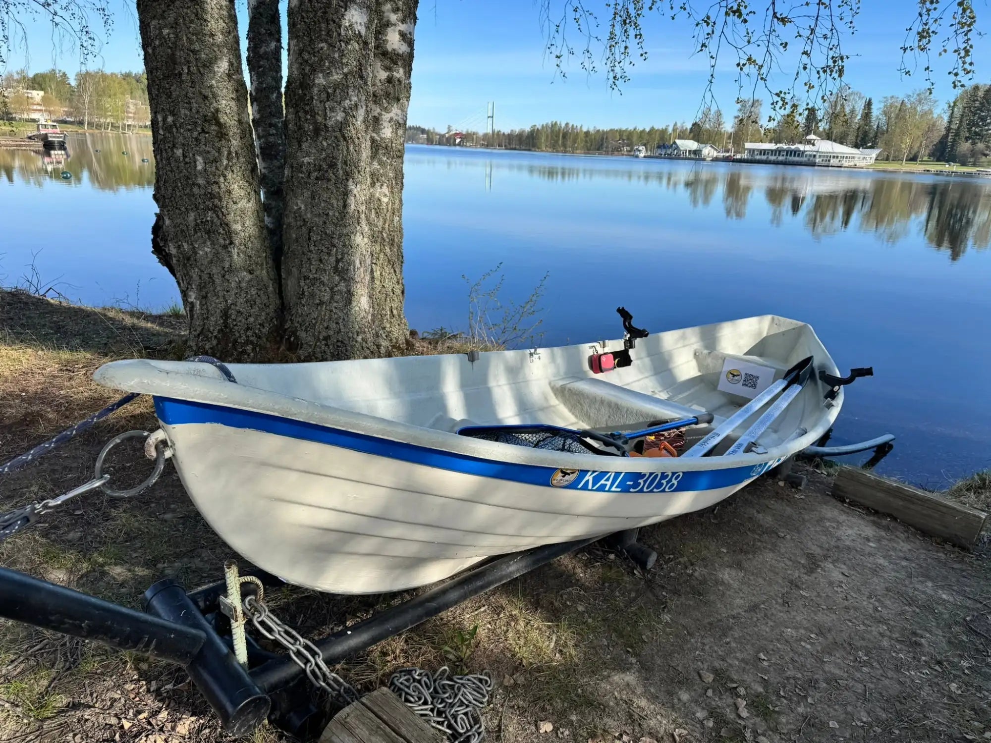 Valkoinen alumiininen soutuvene sinisellä raidalla ja rekisterikilvellä 'kal-3088' kyljessään, levossa perävaunulla rauhallisen järven rannalla puun lähellä.